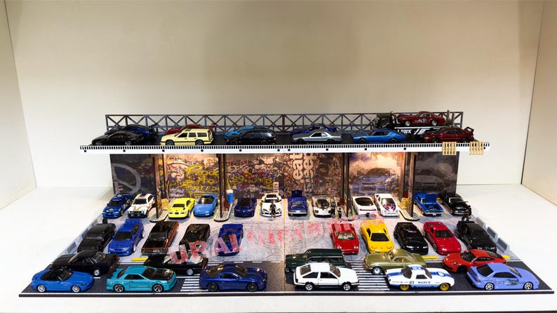 May include: A multi-level parking garage display with a variety of miniature cars in different colors, including red, blue, yellow, black, and white. The cars are arranged in rows on the display, with some cars on the upper level and others on the lower level. The display is made of wood and metal and has a graffiti-style backdrop.