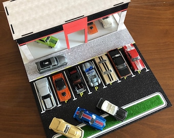 May include: A model car dealership with a white building and a black parking lot. The lot is filled with various model cars, including a black and white police car, a red car, a yellow car, and a blue car. The cars are parked in a row with the word "P" painted on the ground in front of each car.