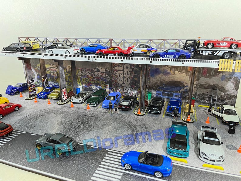 May include: A collection of miniature toy cars in a variety of colors, including red, blue, yellow, and white, are parked in a miniature garage setting. The cars are arranged in rows and include a tow truck with a red car on the back. The garage has a graffiti-style wall with the words "Eat Sleep Repeat" written on it.