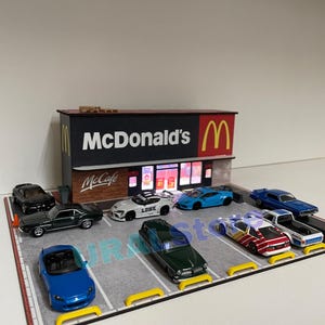 May include: A miniature diorama featuring a McDonald's restaurant with a parking lot filled with toy cars. The McDonald's sign is red and yellow. Various toy cars are parked in the lot, including a blue convertible and a green classic car.