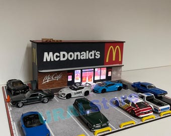Może przedstawiać: Miniaturowa diorama przedstawiająca restaurację McDonald's z parkingiem wypełnionym zabawkami samochodami. Znak McDonald's jest czerwony i żółty. Na parkingu zaparkowanych jest kilka zabawek samochodów, w tym niebieski kabriolet i zielony klasyczny samochód.