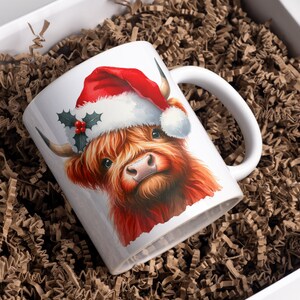 May include: White ceramic mug with a red and white Santa hat on a brown Highland cow. The cow has a red nose and brown fur.