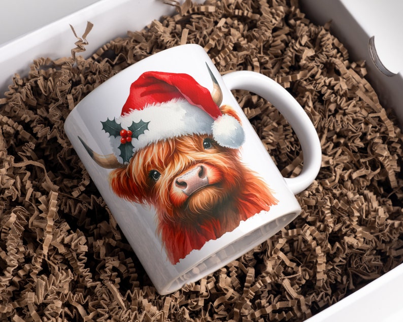May include: White ceramic mug with a red and white Santa hat on a brown Highland cow. The cow has a red nose and brown fur.
