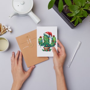 Op de afbeelding: Een groene cactus met een rode kerstmanmuts en versierd met kleurrijke kerstverlichting. De cactus staat in een bruine pot en staat op een wit oppervlak.