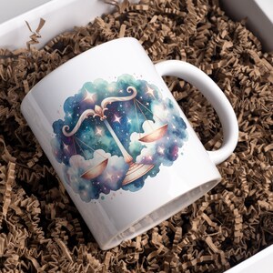 May include: White ceramic mug with a watercolor illustration of the Libra zodiac sign in a galaxy setting. The scales are gold and the background is a blue and purple nebula with stars.