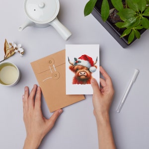 May include: A Christmas card featuring a Highland cow wearing a Santa hat with holly and berries. The cow is red and white with a brown nose and has a white background.