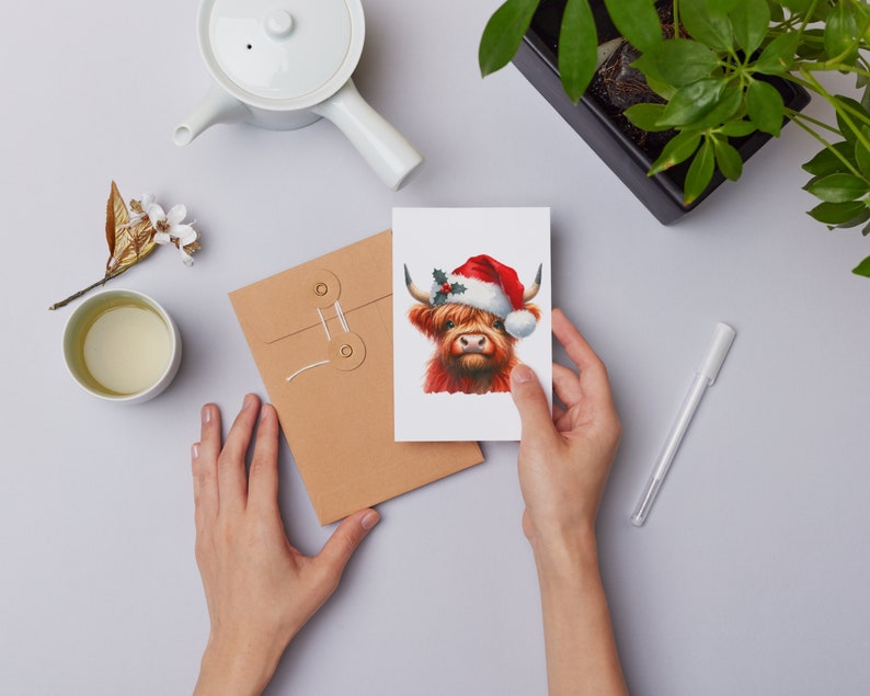 May include: A Christmas card featuring a Highland cow wearing a Santa hat with holly and berries. The cow is red and white with a brown nose and has a white background.