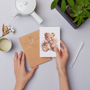 Może przedstawiać: Kartka okolicznościowa z akwarelową ilustracją misia z r&oacute;żową kokardą i kwiatowymi detalami. Kartka trzymana jest w dłoniach, obok brązowej koperty, białego czajniczka i filiżanki herbaty.