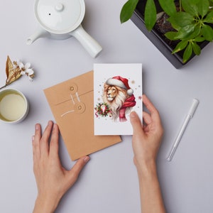 Puede incluir: Una tarjeta de Navidad con un le&oacute;n que lleva un gorro de Pap&aacute; Noel y una bufanda roja. El le&oacute;n est&aacute; rodeado de copos de nieve y acebo.