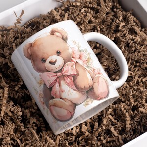 Może przedstawiać: Biały ceramiczny kubek z akwarelową ilustracją misia z r&oacute;żową kokardą i kwiatowymi akcentami. Kubek jest prezentowany w białym pudełku prezentowym z brązowym papierem falistym.