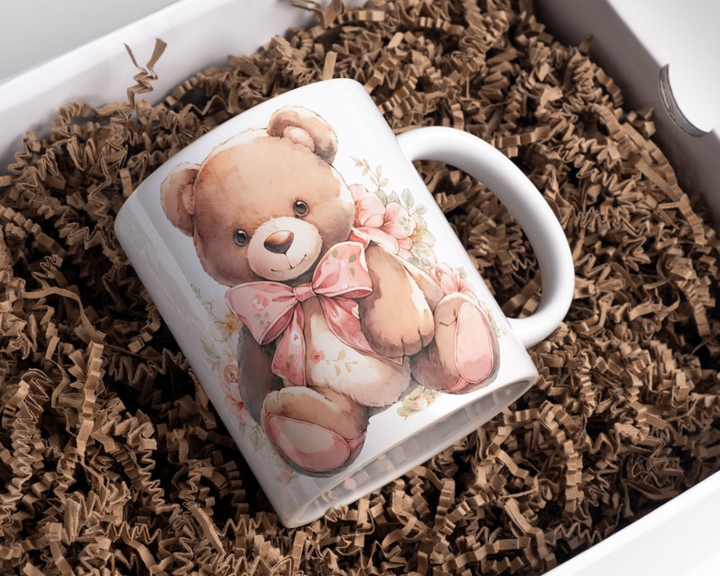 Może przedstawiać: Biały ceramiczny kubek z akwarelową ilustracją misia z r&oacute;żową kokardą i kwiatowymi akcentami. Kubek jest prezentowany w białym pudełku prezentowym z brązowym papierem falistym.