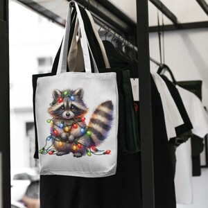 May include: White tote bag featuring a cartoon raccoon adorned with colourful Christmas lights. The raccoon is standing against a white backdrop.