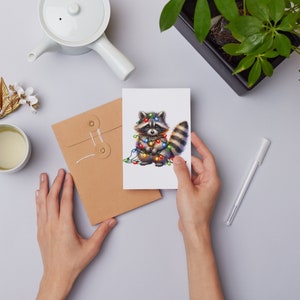 May include: A greeting card featuring a cute cartoon raccoon wearing a string of colourful Christmas lights. The raccoon is standing on a white background.