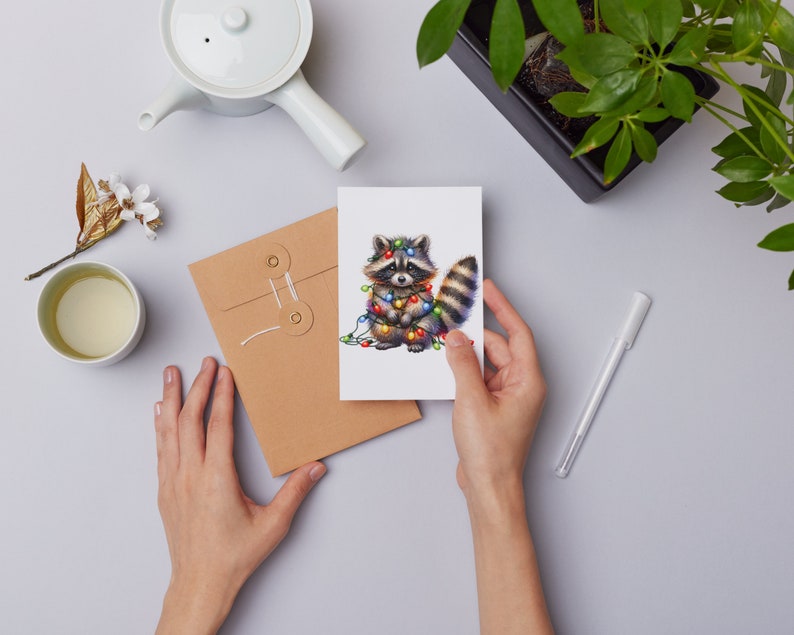 May include: A greeting card featuring a cute cartoon raccoon wearing a string of colourful Christmas lights. The raccoon is standing on a white background.