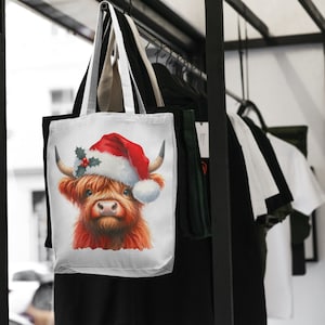 May include: White canvas tote bag with a red and white Christmas hat on a Highland cow. The cow has a red nose and brown eyes. The bag is hanging on a rack with other bags.