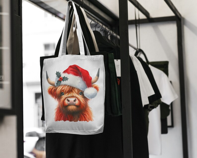 May include: White canvas tote bag with a red and white Christmas hat on a Highland cow. The cow has a red nose and brown eyes. The bag is hanging on a rack with other bags.