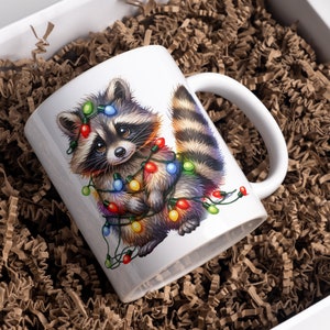 May include: White ceramic mug featuring a colourful illustration of a raccoon wearing Christmas lights. The raccoon is looking at the viewer with a cute expression.