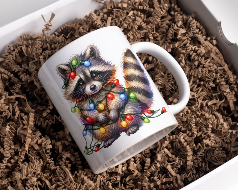 May include: White ceramic mug featuring a colourful illustration of a raccoon wearing Christmas lights. The raccoon is looking at the viewer with a cute expression.