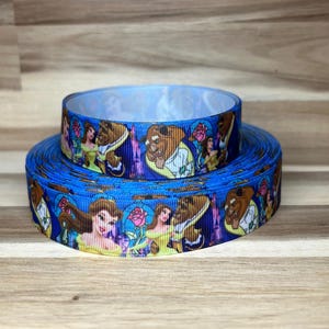 May include: A blue grosgrain ribbon featuring a repeating pattern of Belle and the Beast from Beauty and the Beast, with illustrations of a pink rose in a glass dome. The ribbon is wound into two stacked circular rolls of varying diameters.