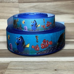 May include: Two stacked rolls of blue grosgrain ribbon featuring a repeating pattern of animated sea creatures, including a blue tang fish, orange clownfish, a sea turtle, and a small purple jellyfish set against an underwater coral reef background.