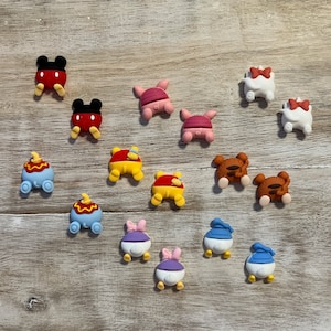 May include: A collection of colorful cartoon character charms, including Mickey Mouse, Piglet, Winnie the Pooh, Tigger, Daisy Duck, and Donald Duck. The charms are in various colors, such as red, yellow, blue, and white, and are arranged on a light wood-grain surface.