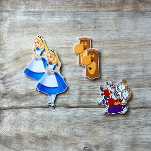 Op de afbeelding: Alice in Wonderland-taarttoppers met twee Alice-figuren in blauwe en witte jurken, twee bruine houten figuren met gezichten en een groep van drie witte konijnen met een zakhorloge.
