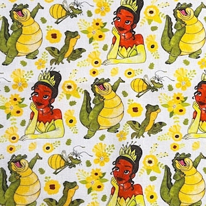 May include: White fabric featuring a repeating pattern of a princess, an alligator, a frog, a bee, and yellow flowers. The princess is wearing a crown and a yellow dress. The alligator is yellow and green. The bee is yellow and black.