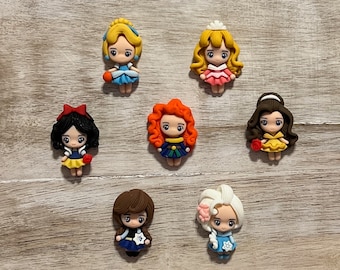 May include: Seven small, colourful, cartoon-style figurines of princesses. Each figurine is about 5 cm tall and has a different hairstyle, dress, and accessories. The figurines are arranged in a circle on a wooden surface.