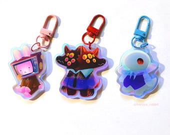 May include: Three holographic acrylic keychains with cartoon character designs. One features a rabbit with a TV head, another a two-headed dog, and the third a ghost-like figure. Each keychain has a different colored clasp: pink, red, and blue.