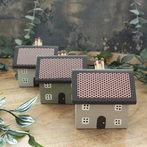 May include: Three wooden matchbox holders shaped like houses. The houses are painted in shades of grey and green with brown roofs. The roofs have a honeycomb pattern. Each house has a small door and windows.