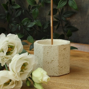 May include: A white ceramic incense holder with a hexagonal shape and a speckled texture. The holder has a small opening at the top for incense sticks.