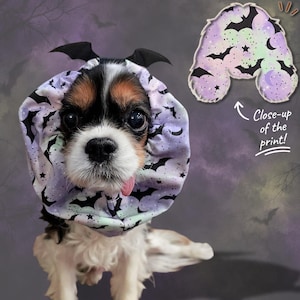 May include: A dog wearing a ruffled collar featuring a pastel purple, green, and white pattern of black bats, stars, and moons. The collar has small black bat wings attached to the top. A separate swatch shows the fabric print with the text Close-up of the print!