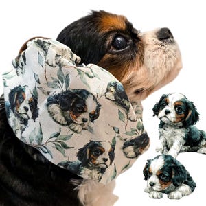 May include: A dog with a patterned fabric accessory featuring puppy portraits and foliage. The dog has a tricolour coat of black, white, and brown. Two puppy figurines are also visible. The accessory is wrapped around the dog's neck.