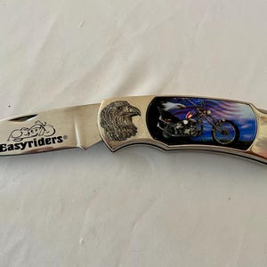 May include: A silver folding knife with the text "Easyriders" on the blade. The handle features an eagle head and a motorcycle design against a backdrop of the American flag. The knife is closed.