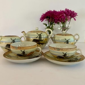 May include: A vintage tea set featuring a teapot, sugar bowl, creamer, and two teacups with saucers. Each piece is painted with a landscape scene of a windmill. The set has a cream and tan color scheme with gold trim. A bouquet of pink flowers is in the background.