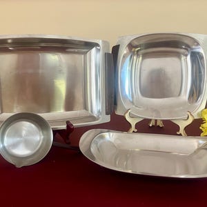 May include: A collection of vintage stainless steel serving dishes. Includes a rectangular tray with wooden handles, a square bowl, a small round dish, and an oval serving dish. The set is displayed on a red surface.