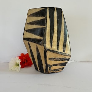 May include: A decorative, geometric vase with a black and tan pattern. The vase has a unique, angular shape and is decorated with black stripes and triangles against a tan background. The vase is a decorative item.