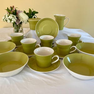 May include: A complete vintage lime green and white ceramic tea set, including cups, saucers, bowls, a sugar bowl, a creamer, and serving dishes. A vase of flowers is in the background. The set is arranged on a white tablecloth.