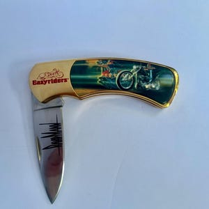 May include: A gold-colored pocket knife with a stainless steel blade. The knife features the text "Easyriders" in red and a motorcycle scene on the handle. The blade is partially open, displaying a black graphic.