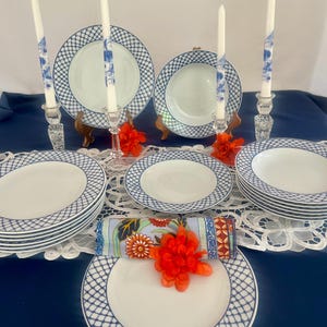 May include: A dinnerware set featuring white plates and bowls with a blue lattice pattern on the rims. The set includes stacks of plates, bowls, four tall white taper candles with blue floral designs in glass holders, and a floral patterned napkin.