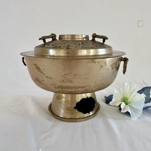 以下が含まれることがあります： 蓋付きのヴィンテージ真鍮製調理鍋。鍋は丸い本体とハンドル、装飾的な蓋が付いています。鍋は開口部のある台座に置かれています。鍋は温かみのある金色です。