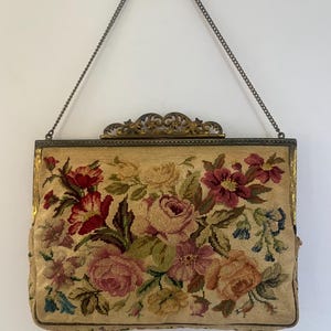 Puede incluir: Bolso de mano vintage de tapiz con un diseño floral en tonos rojos, rosas y amarillos. El bolso rectangular tiene un marco de metal y un asa de cadena. El estampado floral está sobre un fondo beige.