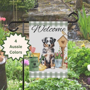 May include: A garden flag with a green and white checkered background. The flag features a blue merle Australian Shepherd puppy sitting in a garden setting with a watering can, flowers, and a birdhouse. The text "Welcome" is at the top of the flag and "Maggie's Garden" is at the bottom.