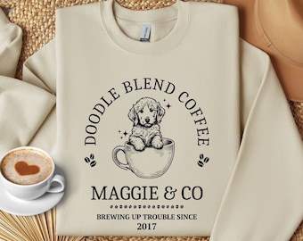 May include: A beige sweatshirt with a black graphic design featuring a doodle dog in a coffee cup. The text reads "Doodle Blend Coffee Maggie & Co Brewing Up Trouble Since 2017".