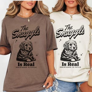 May include: Two t-shirts, one espresso and one ivory, featuring a graphic of a puppy with the text "The Snuggle Is Real, Resistance Is Futile." The shirts are short-sleeved and have a relaxed fit. The design is in black.