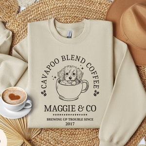 May include: A beige sweatshirt with a black graphic design of a dog in a coffee cup. The text on the graphic reads "Cavapoo Blend Coffee, Maggie & Co, Brewing Up Trouble Since 2017".