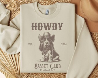 May include: Sand-colored sweatshirt with a graphic of a Basset Hound wearing a cowboy hat and bandana. The text "HOWDY" is above the dog, with "BASSET CLUB Portland, ME" below. The sweatshirt has a crew neck.