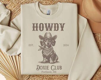 May include: Sand-colored sweatshirt with a graphic of a dachshund wearing a cowboy hat and bandana. The text reads "HOWDY" above the dog and "DOXIE CLUB Portland, ME" below. The sweatshirt is surrounded by woven decor and a brown hat.