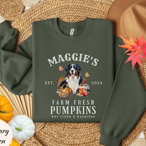 May include: Military green sweatshirt with the text "Maggie's Farm Fresh Pumpkins" and a Bernese Mountain Dog illustration. The design includes pumpkins, fall leaves, and the year 2024. A fall-themed apparel item.