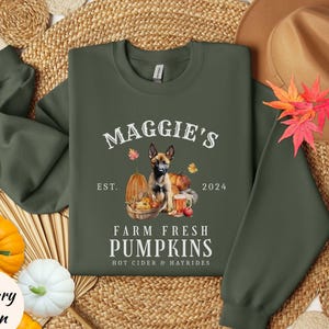 May include: Military green sweatshirt with the text "Maggie's Farm Fresh Pumpkins" and a dog graphic. The design includes pumpkins, fall leaves, and the year 2024. The sweatshirt is surrounded by fall-themed decorations.